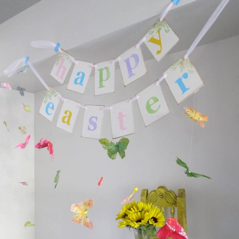 May include: A pastel-colored banner with the words "happy easter" hanging from a white ceiling. The banner is decorated with small, colorful butterflies.