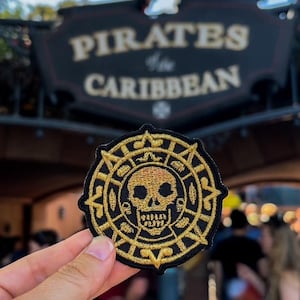 May include: A gold embroidered patch with a skull and crossbones design, surrounded by a compass rose. The patch is held in front of a sign that reads "Pirates of the Caribbean".