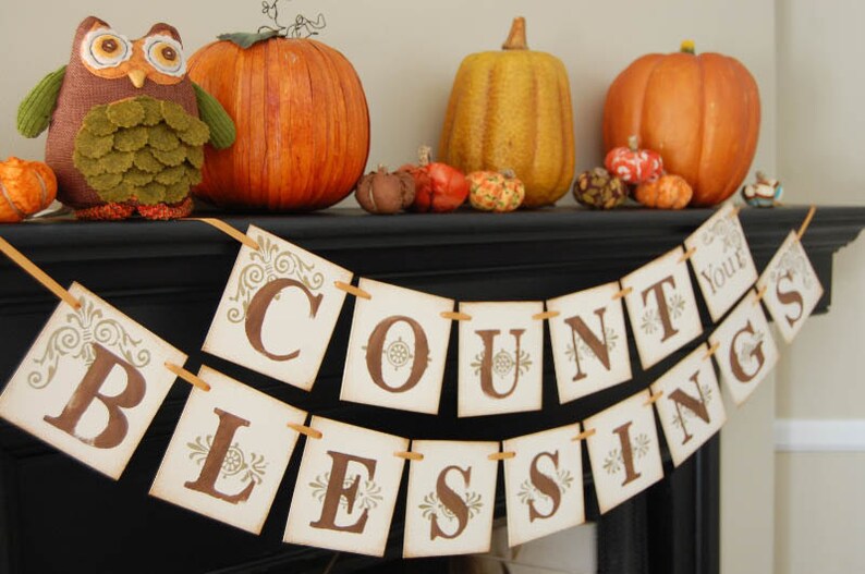 May include: A brown and green owl figurine sits on a black mantel with a banner that reads "Count Your Blessings". The banner is made of white paper with brown lettering and gold accents. There are also several small pumpkins and gourds on the mantel.