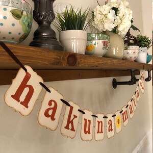 May include: A banner with the phrase "Hakuna Matata" in red letters on white paper with a yellow sunflower in the middle of the banner. The banner is hanging from a brown wooden shelf with a black pipe.