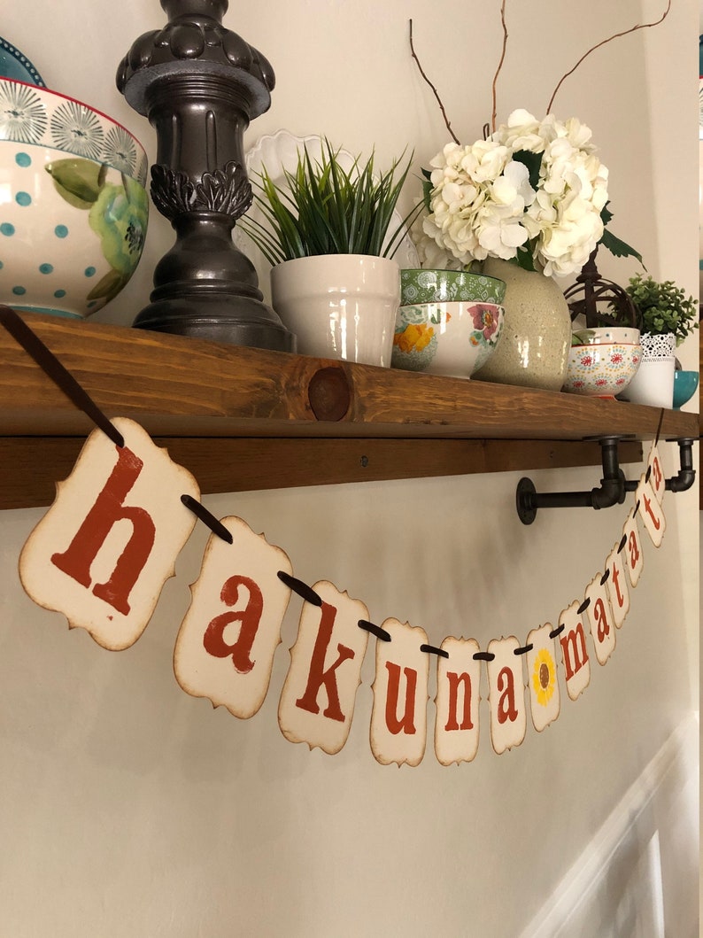 May include: A banner with the phrase "Hakuna Matata" in red letters on white paper with a yellow sunflower in the middle of the banner. The banner is hanging from a brown wooden shelf with a black pipe.