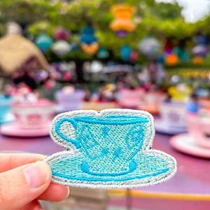 May include: A blue embroidered teacup and saucer patch. The patch is a whimsical design with a white outline and a light blue background.