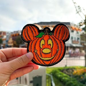 May include: A Halloween-themed embroidered patch featuring Mickey Mouse as a pumpkin. The patch is orange and black with a green stem and Mickey's signature smile.