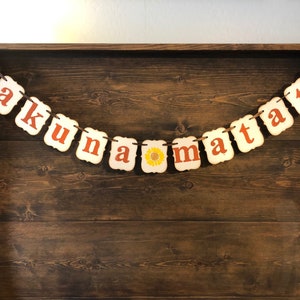 May include: A white banner with the phrase "Hakuna Matata" in orange letters. The banner is strung with twine and has a small sunflower graphic between the words "na" and "ma".
