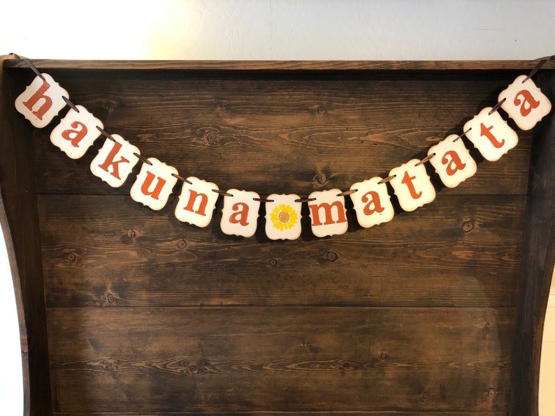 May include: A white banner with the phrase "Hakuna Matata" in orange letters. The banner is strung with twine and has a small sunflower graphic between the words "na" and "ma".