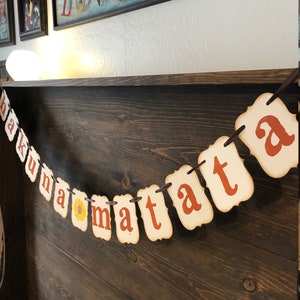 May include: A banner with the phrase "Hakuna Matata" in orange letters on a cream background. The banner is strung with twine and has a sunflower graphic between the words "na" and "ma".