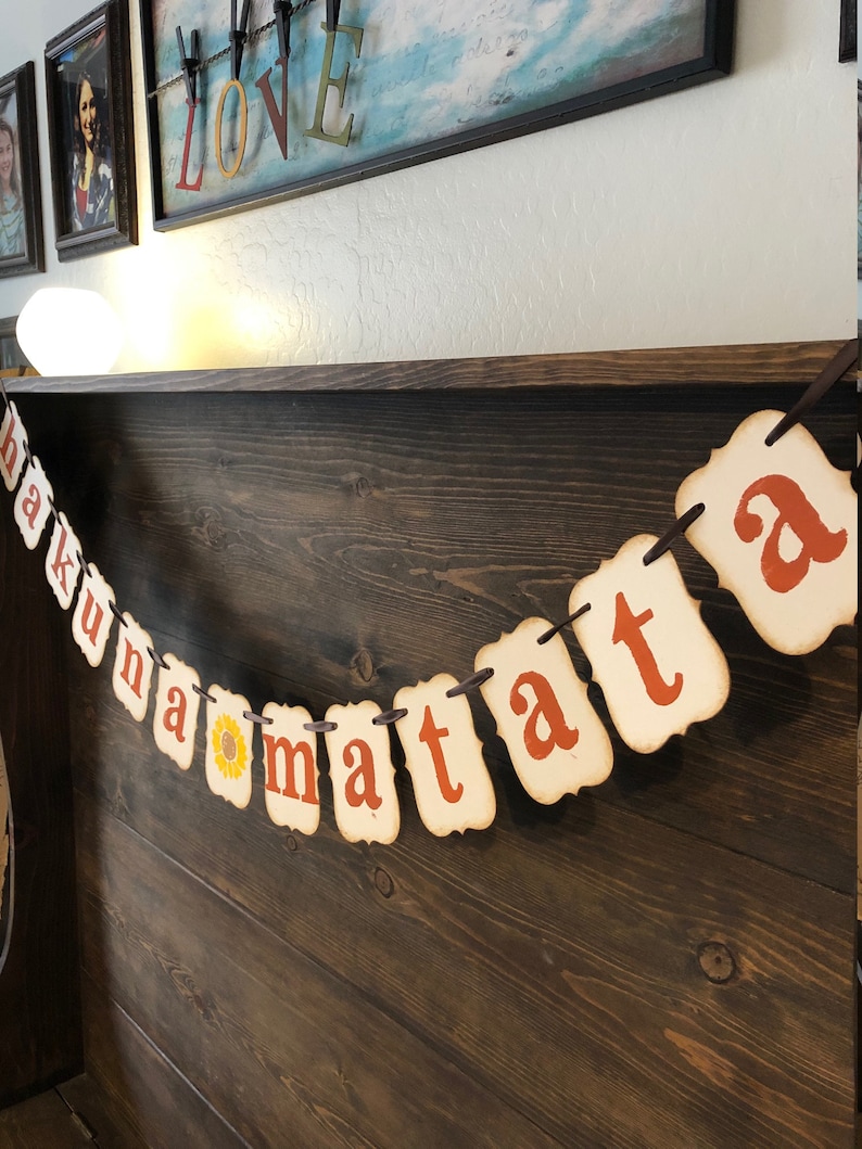 May include: A banner with the phrase "Hakuna Matata" in orange letters on a cream background. The banner is strung with twine and has a sunflower graphic between the words "na" and "ma".