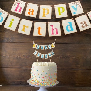 May include: A white cake decorated with colorful sprinkles and a banner that says "Happy Birthday" in colorful letters. A smaller banner on top of the cake says "Baby Landen".