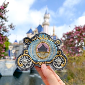 May include: A blue and gold embroidered patch of Cinderella's carriage with a crown on top. The patch is being held in front of a blurred image of Cinderella's castle.