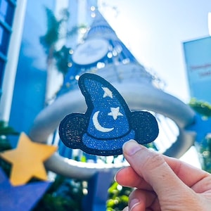 May include: A blue embroidered patch of Mickey Mouse's Sorcerer's Hat with a white crescent moon and stars. The patch is being held in front of a blue and yellow star.