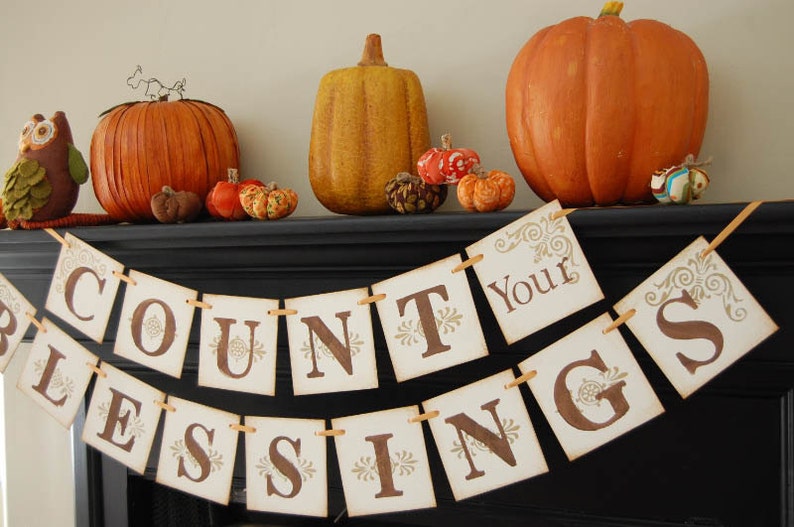 May include: A Thanksgiving banner with the words "Count Your Blessings" in brown letters on a white background. The banner is decorated with fall-themed decorations, including pumpkins, gourds, and pinecones.