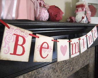 May include: A Valentine's Day banner with red and pink paper flags that spell out "Be mine". The flags are strung together with red ribbon and are hanging on a black mantel.