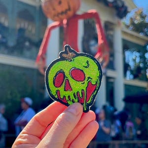 May include: A green apple with a red skull and dripping green and red icing. The apple is a Halloween-themed patch.