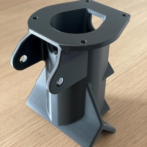 Puede incluir: Un componente gris oscuro impreso en 3D con una parte superior rectangular con cuatro agujeros y una abertura central. El componente tiene un cuerpo cilíndrico con soportes angulados y dos brazos laterales con agujeros. El objeto está sobre una superficie de madera.