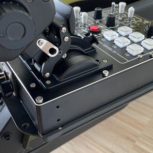 May include: A black flight simulator control panel with various buttons, switches, and a joystick. The panel has a silver trim and is mounted on a black frame. A black cable is connected to the side.