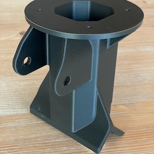 May include: A black plastic 3D printed adapter with a hexagonal opening and a flat base. The adapter has a side arm with a hole and a curved bottom.