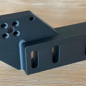 May include: A black plastic 3D printed part with a circular cutout and five holes in the centre. The part has a rectangular extension with two rectangular cutouts.