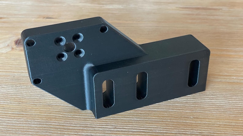 May include: A black plastic 3D printed part with a circular cutout and five holes in the centre. The part has a rectangular extension with two rectangular cutouts.