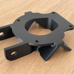 Puede incluir: Una pieza negra impresa en 3D con una abertura circular y dos brazos que se extienden hacia afuera. La pieza tiene varios agujeros pequeños en la superficie.