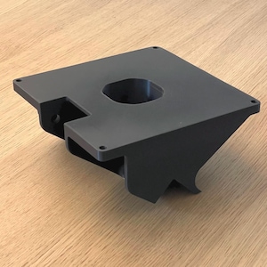 Op de afbeelding: Een zwarte, 3D-geprinte basis van plastic met een ronde opening in het midden. De basis heeft een uniek ontwerp met een gebogen, hoekige vorm.
