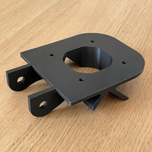 Può includere: Pezzo in plastica nera stampato in 3D con un grande foro circolare e diversi fori più piccoli. Il pezzo ha un design unico con una base curva e due bracci che si estendono.