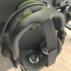 Op de afbeelding: Een zwarte VR-headset met een groene band hangt aan een zwarte stoel. De headset heeft een zwarte band en een zwart hoofdband. De headset hangt aan een zwarte stoel met een groene band.
