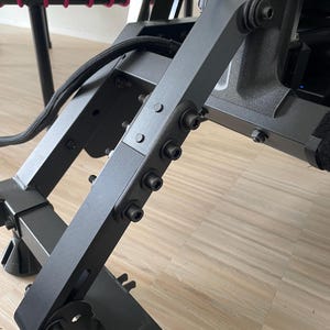 May include: Close-up of a black metal frame with multiple bolts and a black cable. The frame is attached to a larger black metal structure.