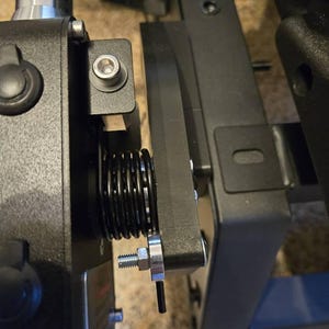 May include: Close-up of a black metal mechanism with a spring and a silver screw. The mechanism is attached to a black metal frame.