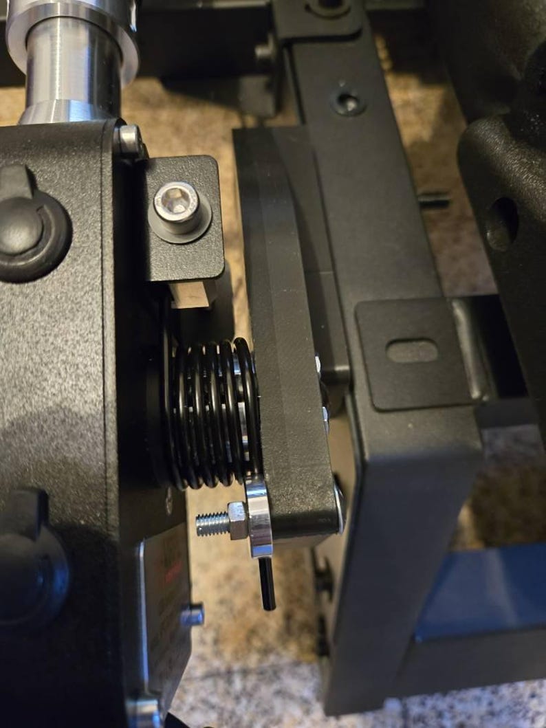 May include: Close-up of a black metal mechanism with a spring and a silver screw. The mechanism is attached to a black metal frame.