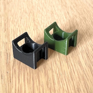 Op de afbeelding: Twee kleine, 3D-geprinte houders. Eén is zwart, de andere is olijfgroen. Elk heeft een vierkante basis met een cirkelvormige opening en een rechthoekige bovenkant met een handvat. De houders zijn ontworpen om een klein object vast te houden.
