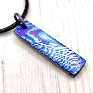 Könnte beinhalten: Ein blauer und violetter, gemusterter Metallanhänger an einer schwarzen Schnurkette. Der Anhänger ist rechteckig und hat ein wellenförmiges Muster.