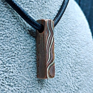 May include: A rectangular pendant featuring a layered mokume gane pattern of copper, brass, and nickel silver tones. The pendant is suspended from a simple black cord necklace, showcasing a vertical orientation with a hole at the top for the cord.