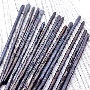 May include: A collection of fourteen long, slender metal rods with a dark, hammered finish. Each rod features a rounded tip and a textured, uneven surface created by the hammering process, displaying variations in colour from charcoal to bronze.