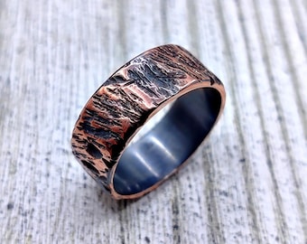May include: A wide band ring featuring a textured, rugged exterior with a metallic copper finish and dark, recessed detailing. The interior of the band is smooth and polished with a dark, gunmetal grey finish, creating a distinct contrast.
