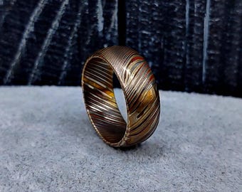 May include: A ring featuring a complex, multi-layered pattern of metallic bands in shades of copper, bronze, and dark steel. The surface displays a swirling, wood-grain-like texture created by the alternating layers of metal, forming a continuous band.