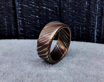 May include: A ring featuring a layered mokume gane pattern with alternating bands of copper, brass, and silver tones. The metal surface displays a continuous, swirling, diagonal striped texture that wraps around the entire circumference of the band.