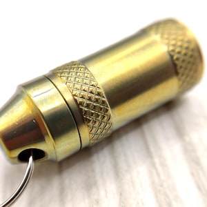 May include: A gold-toned metal keychain with a small, cylindrical container attached. The container has a textured, diamond-shaped pattern and a key ring.