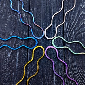 May include: Six wavy hair clips in different colors, including blue, purple, green, yellow, and silver, arranged in a starburst pattern on a dark wood background.