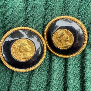 May include: A pair of round, gold-toned clip-on earrings. Each earring features a black center with a gold coin-like design and a gold rope-like border. The coin design has a profile of a person.