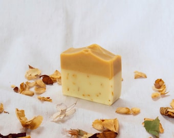 Puede incluir: Una pastilla de jabón rectangular con un diseño de dos tonos, con una capa superior de color bronceado sólido y una capa inferior de color blanco crema que contiene pequeñas partículas botánicas incrustadas, rodeada de pétalos y hojas secas.