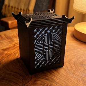 May include: A square black lantern with a roof-like top and intricate geometric cutouts on the sides. The lantern emits a soft white light from within, highlighting the detailed patterns on its panels and the textured surface of the top.