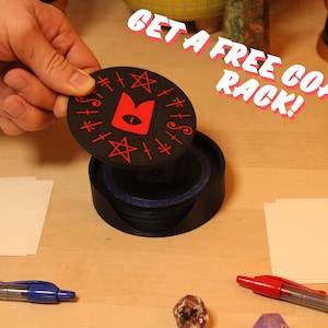 May include: A black coaster rack with a red cat design on the top coaster. The text "GET A FREE COASTER RACK!" is written in red letters on a white background.