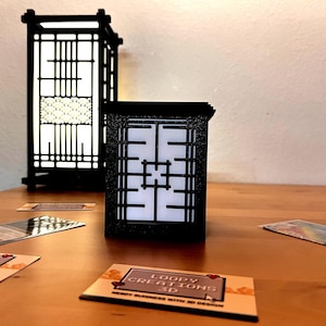 May include: Two black, geometric lanterns with white interiors sit on a wooden surface. The lanterns have a traditional Japanese design. Business cards with the text "Loopy Creations 3D" are scattered around the lanterns.