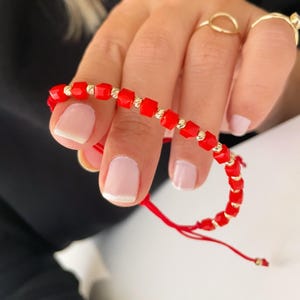 Könnte beinhalten: Ein rotes Perlenarmband mit goldenen Akzenten. Das Armband besteht aus kleinen, quadratischen Perlen und ist mit einer roten Schnur gebunden.