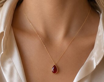 May include: A delicate gold chain necklace featuring a single teardrop-shaped pendant with a deep red faceted gemstone. The pendant is encased in a thin gold setting, suspended from a fine, polished gold link chain that rests against the collarbone.