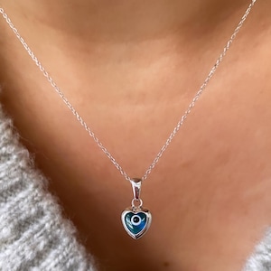 Może przedstawiać: Srebrny naszyjnik z łańcuszkiem z sercowatym wisiorem z niebieskim amuletem w kształcie oka. Wisiorek jest otoczony srebrną obwódką.