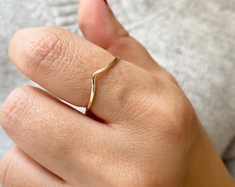 May include: A delicate gold-coloured ring in a V-shape design. The ring is worn on a finger, showcasing its minimalist and elegant style. The ring's smooth surface reflects light, highlighting its polished finish. The jewellery piece is a simple yet stylish accessory.