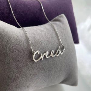Può includere: Una collana color argento con il nome "Creed" in corsivo. La collana è esposta su un cuscino grigio con uno sfondo viola.