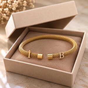 Op de afbeelding: Een goudkleurige mesh armband met twee gepolijste cilindrische eindkappen en twee bevestigde metalen letters, een hoofdletter I en een hoofdletter B, rustend in een beige rechthoekige geschenkdoos met een zachte stoffen voering.