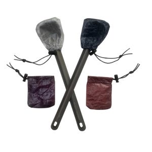 May include: Two titanium camp spoons with protective covers and drawstring pouches. One spoon has a silver cover, the other a navy blue cover. The pouches are purple and maroon. The spoons are crossed against a white background.